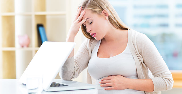 tired pregnant woman sits at desk with eyes closed and hand on forehead and stomach