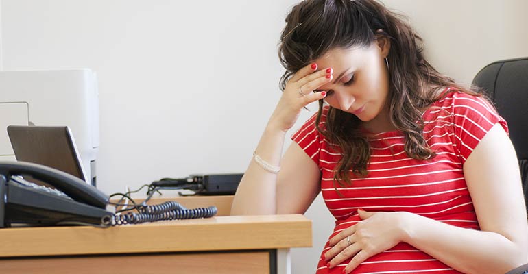 Pregnant woman sitting at desk with head in right and and left hand over stomach