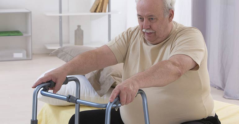 elderly obese man sitting on bed with hands on walker