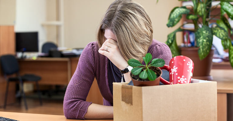 woman sits at desk with head down and box of belongings