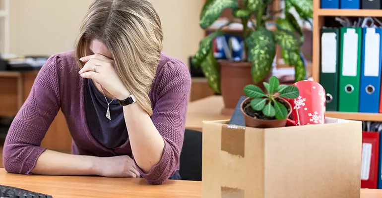 worried woman sitting at desk with box of belongings looking down